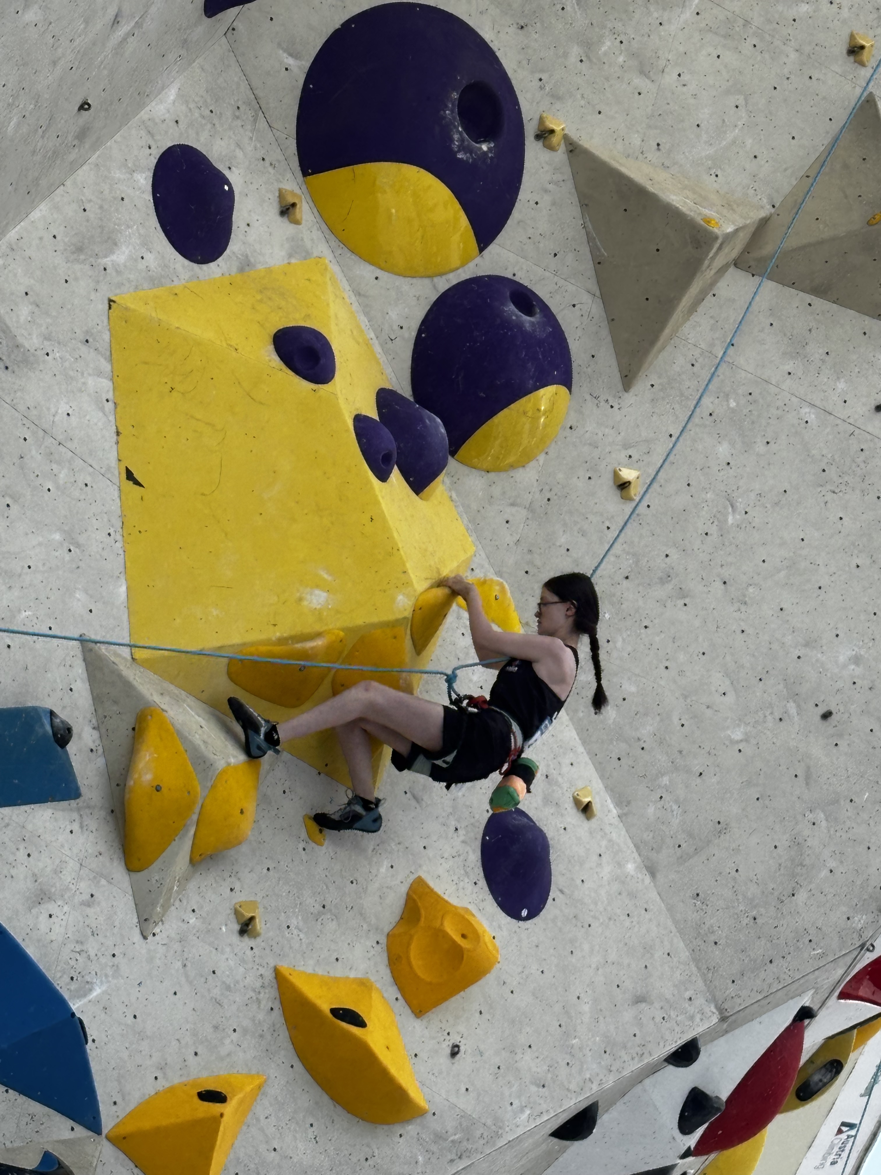 Jackie is climbing on an overhung route with yellow, purple, and blue holds. Her leg right leg is bent on a hold and her left heel is stretched out onto a hold slightly further away.