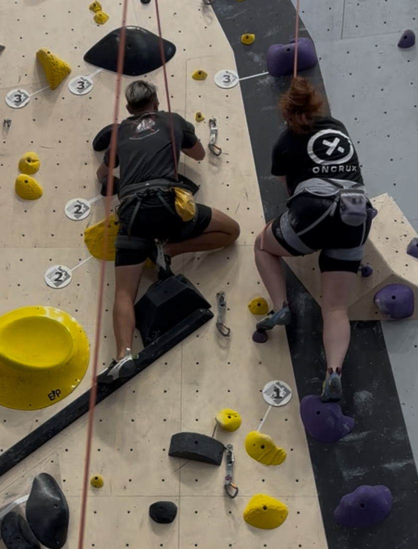 Matt is climbing a black route on the left side of Paige, who is climbing a purple route right next to him. Matt has his right leg bent in a deep squat and his left leg lower down on a longer hold. Paige has her left foot on a hold that’s level with her right knee, and her right legis further down on a bigger hold.