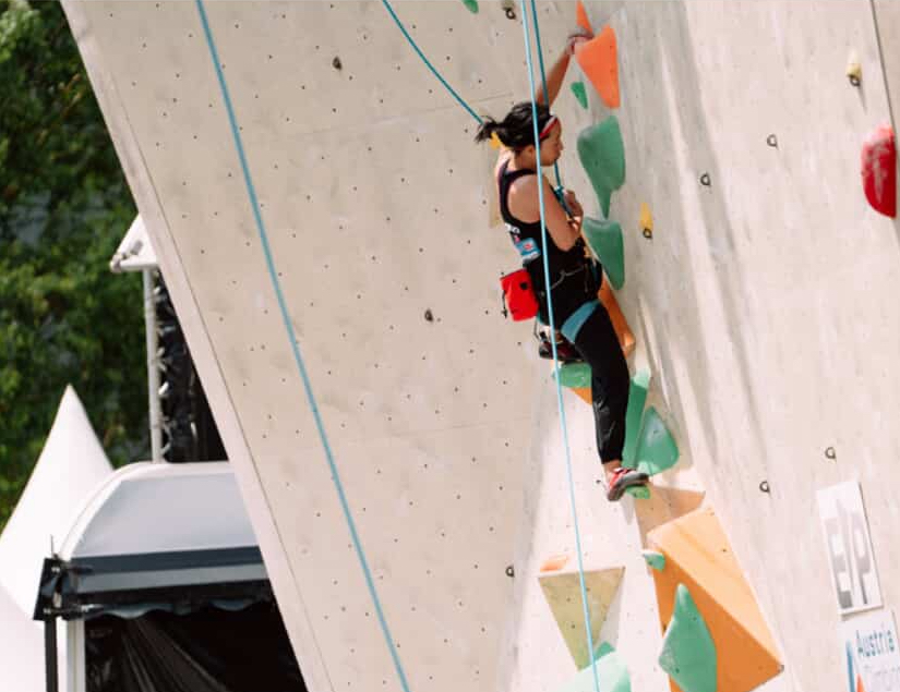 Tiffany climbs a route with mint green and orange holds. Her left heel is on a hold out to the left and her left hand is holding on to a hold above her head. Her right hand is close to her body and she’s looking at the wall as she decides what to do next.